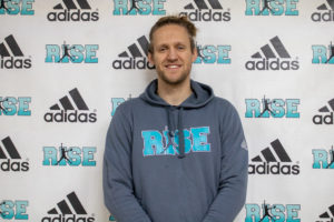 A man smiling in a blue hoodie with a branded backdrop.